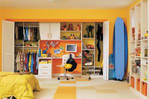 A reach in kid's closet with custom shelves, hanging rods, metal basket and work desk in a wood grain finish with bright accents by California Closets.