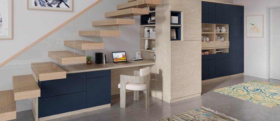 Oficina en casa con acabado en madera veteada clara con detalles en azul intenso, despensa y armarios de almacenamiento debajo de las escaleras de California Closets