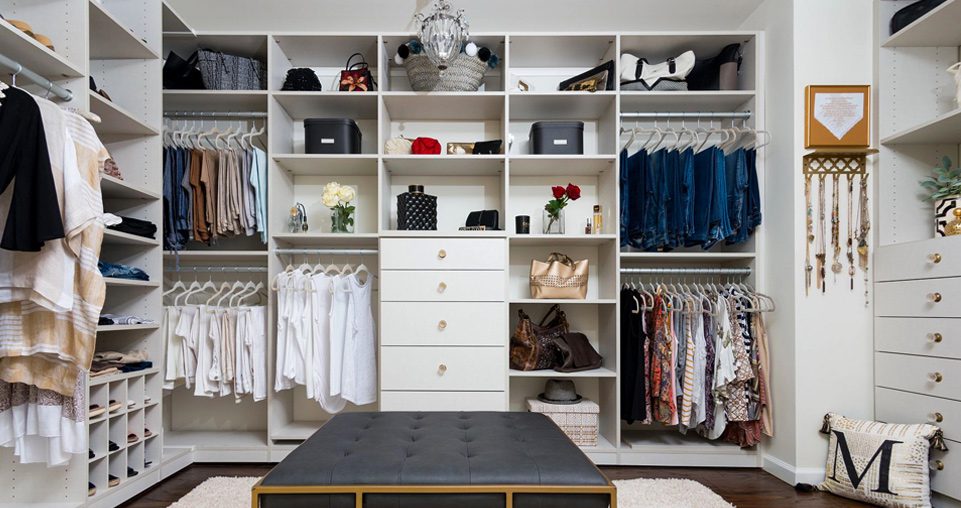 White custom walk in closet with blue ottamin, custom shelving, hanging racks, shoe storage, and cubbies Designed by California Closets.