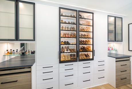 His and hers walk in closet design with LED lighting to illuminate shoe storage through glass doors created by California Closets