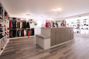 A walk in closet design with a boutique feel is designed with custom cabinets, drawers, LED lighting, shoe storage and a center island created by California Closets