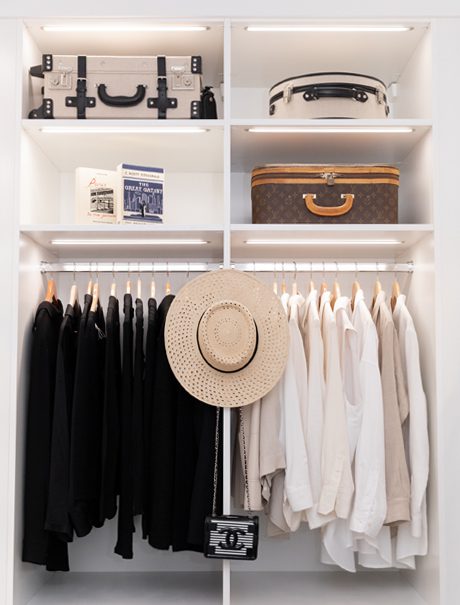 Closet shelving and hanging clothes poles under LED lighting in a white wood grain finish by California Closets
