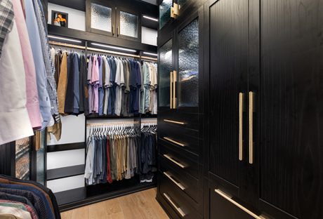His side of the his and hers closet in a dark wood grain finish with gold hardware, showcasing custom shelving, a built in dresser and led lighting by California Closets