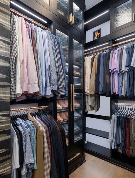 Walk in closet designed for his side in dark wood grain finish with custom shoe storage behind cabinet doors with glass inserts created by California Closets